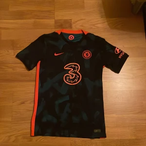 Chelsea FC bortatröja Lukaku #9 Nike - Chelsea FC bortatröja med Lukaku #9 på ryggen. Tröjan är svart med blått kamouflagemönster och har orange detaljer längs sidorna och kragen. Nike-logga och klubbmärke på bröstet. Tillverkad i Dri-FIT-material för bra ventilation.