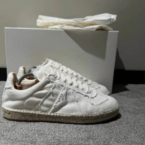 Maison margiela Espadrillos Vita sneakers i canvas med repdetalj - Säljer ett par vita sneakers i canvas med råa kanter och repdetalj runt sulan. Skorna har snörning och en platt sula, perfekt för en avslappnad stil. Klassisk design med modern twist.perfekt för sommarn och retail är 7000 köpta i Italien.