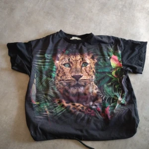 Svart t-shirt med leopardtryck - Cool svart t-shirt med stort leopardmotiv framtill och gröna blad runtom. T-shirten har korta ärmar och en avslappnad passform, perfekt för dig som gillar djurmotiv.