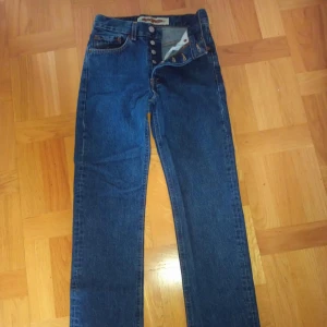 Blå jeansbyxor från Crocker Jeans Company - Klassiska blå jeansbyxor från Crocker Jeans Company med raka ben och hög midja. Byxorna har knäppning med fyra silverfärgade knappar och tydliga kontrastsömmar. Två bakfickor och två framfickor. Perfekta för en tidlös denimlook.  Måten på byxorna är W:26 L:32 . 