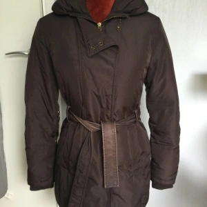 Plommonfärgad Dunjacka Jacka Dun Strl: S MNG - Säljer en brun parkajacka med huva och bälte i midjan. Jackan har långa ärmar, dragkedja framtill, knappar och snygga detaljer med dragkedjor vid ärmsluten. Perfekt för kyligare dagar. Plommonfärgad dunjacka Längd: 72 cm Armhåla till armhåla: 46 cm Dragkedja och tryckknappar Mudd på ärmslut. Katter finns i hemmet! 