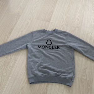 Grå sweatshirt från Moncler - Säljer en stilren grå sweatshirt från Moncler med svart logotyp och text på bröstet. Tröjan har rund halsringning och långa ärmar. Perfekt för en avslappnad och trendig look.