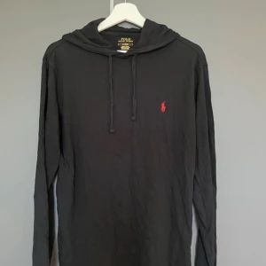 Svart hoodie från Polo Ralph Lauren - Svart hoodie från Polo Ralph Lauren med klassisk huva och dragsko. Tröjan har långa ärmar och en liten röd broderad logga på bröstet. Perfekt för en avslappnad stil.