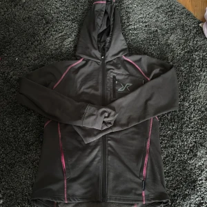 Svart fleecejacka med huva från RevolutionRace - Mysig svart fleecejacka från RevolutionRace med huva och dragkedja. Jackan har rosa detaljer, praktiska fickor med dragkedja och en bröstficka. Perfekt för utomhusaktiviteter och träning.