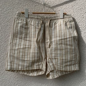 Beige och vita randiga shorts från H&M - Randiga shorts i beige och vitt från H&M med snörning i midjan. Tillverkade i en mix av linne och bomull för en luftig känsla. Shortsen har normal passform och fickor både fram och bak.
