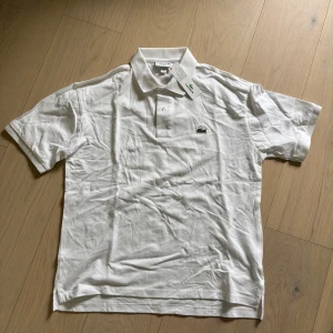 Vit pikétröja från Lacoste - Klassisk vit pikétröja från Lacoste med korta ärmar och krage. Tröjan har den ikoniska broderade krokodilloggan på bröstet och knäppning vid halsen. Perfekt för en stilren och sportig look.