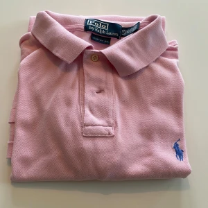 Rosa pikétröja från Polo Ralph Lauren - Säljer en klassisk rosa pikétröja från Polo Ralph Lauren med blå broderad logga på bröstet. Tröjan har krage, knappar vid halsen och är kortärmad. Perfekt för en stilren och avslappnad look. Storlek L
