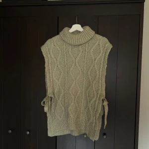 Beige stickad väst med polokrage från Mazing - Säljer en beige stickad väst från Mazing med polokrage och knytband i sidorna i storlek S/M. Västen har ett snyggt kabelstickat mönster och är perfekt att styla över en skjorta eller tröja. Oversized passform för en trendig look.