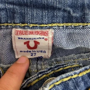 Blå jeans från True Religion - Säljer ett par klassiska blå jeans från True Religion med kontrastsömmar och broderade bakfickor. Jeansen har normal passform och är tillverkade i denim med snygga slitningar framtill och bak.