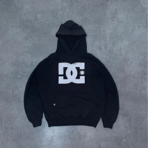 Svart hoodie från DC Shoes - Svart hoodie från DC Shoes med stor vit logga på bröstet. Klassisk huva och magficka framtill. 