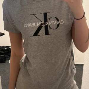 Grå t-shirt från Calvin Klein Jeans - Säljer en klassisk grå t-shirt från Calvin Klein Jeans med stor svart CK-logga och vit text framtill. T-shirten har rund halsringning och korta ärmar. Perfekt till jeans eller shorts för en enkel och stilren look.