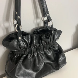 Svart handväska med rynkade detaljer - Snygg svart handväska i skinnimitation med rynkade detaljer och breda axelband. Väskan har dragkedja upptill och rymligt innerfack. Perfekt för dig som vill ha en stilren accessoar med lite extra attityd.