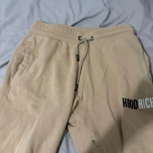 Beige mjukisbyxor från Hoodrich - Säljer ett par beige mjukisbyxor från Hoodrich i storlek M. Byxorna har dragsko i midjan, fickor på sidorna och Hoodrich-logga tryckt på ena benet. Perfekta för en avslappnad stil.