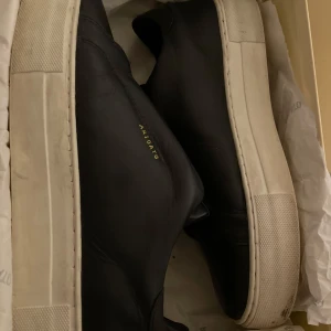 Svarta sneakers från Axel Arigato - Stilrena svarta sneakers från Axel Arigato med vit platt sula och diskret guldfärgad logga på sidan. Skorna är i skinn och har en minimalistisk design utan snörning, vilket ger en clean look.