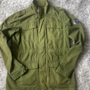 Olivgrön jacka från Fat Moose - Säljer en olivgrön jacka från Fat Moose med fyra fickor framtill och dragkedja. Jackan har hög krage, axelremmar och en dekorativ patch på ena ärmen. Perfekt för dig som gillar stilren och funktionell design.