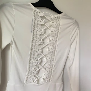 Vit långärmad body med spets och snörning - Säljer en vit långärmad body med snygg spets och snörning i ryggen. Bodyn har rund halsringning och är tillverkad i ett stretchigt material som sitter tight mot kroppen. Perfekt för att skapa en stilren look.