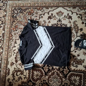 Svart och vit t-shirt från Givenchy Paris - Säljer en svart t-shirt från Givenchy Paris med vita och svarta banddetaljer över bröstet och ärmarna. T-shirten har ett tydligt V-mönster framtill och logga på banden. Klassisk passform och korta ärmar. Perfekt för dig som gillar streetwear.