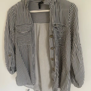 Randig skjorta från Vero Moda - Säljer en snygg vit och blå randig skjorta från Vero Moda i storlek S. Skjortan har långa ärmar, knappar framtill och två bröstfickor. Perfekt till jeans eller kjol för en avslappnad look.
