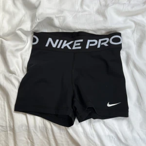 Svarta träningstights shorts från Nike Pro - Svarta tighta shorts från Nike Pro med bred resår i midjan och vit Nike-logga på benet. Perfekta för träning eller sport. Tillverkade i stretchigt syntetmaterial för maximal rörelsefrihet.