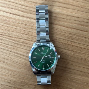 Grön klocka med silverlänk från Omax - Stilren herrklocka från Omax med grön urtavla, datumvisning och silverfärgad länk i metall. Klockan har tydliga indexmarkeringar och kvartsverk. Perfekt accessoar för dig som gillar klassisk design med en modern touch.