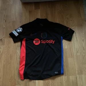 FC Barcelona svart fotbollströja med Spotify sponsor - Säljer en svart FC Barcelona fotbollströja med röda och blå detaljer, Nike-logga och Spotify som sponsor på bröstet. Tröjan har korta ärmar, broderat klubbmärke och UEFA Foundation-märke på ärmen. UNHCR-logga på ryggen.