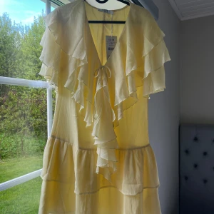 Ljusgul volangklänning  - Supersöt ljusgul klänning från NA-KD i storlek 38 med flera lager volanger och v-ringning med knyt framtill. Klänningen har korta ärmar och är tillverkad i ett lätt, luftigt material som ger en romantisk känsla. Helt ny med prislapp kvar! Perfekt för sommarens alla tillfällen.