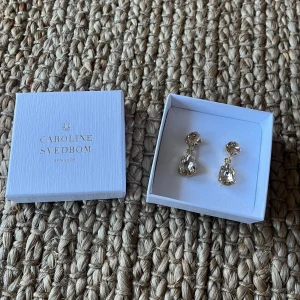 Örhängen från Caroline Svedbom - Helt oanvända! Säljer ett par eleganta örhängen från Caroline Svedbom med stora, gnistrande stenar i droppform och rund form i färgen Silk. Nypris 895kr.