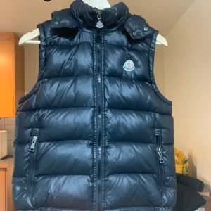 Svart dunväst från Moncler - Snygg svart dunväst från Moncler med hög krage och avtagbar huva. Västen har två fickor med dragkedja framtill och klassisk Moncler-logga på bröstet. Perfekt för lager-på-lager och håller dig varm under kyliga dagar.