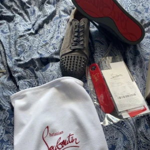 Gråa sneakers med nitar från Christian Louboutin - Säljer ett par gråa sneakers från Christian Louboutin med gråa nitar på tån och den klassiska röda sulan. Skorna har snörning och är tillverkade i mocka. Kommer med dustbag och skohorn. Kvitto följer med 
