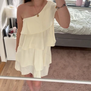 Ljusgul omlottklänning med volanger - Säljer en slutsåld och söt ljusgul klänning med volanger och oneshoulder-design. Klänningen har flera lager och en luftig känsla, perfekt för sommarens tillfällen. Den är kort och har en elegant, asymmetrisk axel. Den är aldrig använd, i storlek 170/S från Gina Tricot!