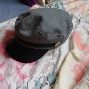 skepparmössa - grå skepparmössa med svart, rundad skärm och band. Mössan har metallknappar på sidorna. 