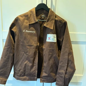 Tyler the creator jacka - Säljer en brun jacka från Golf Wang med broderad text 'T. Baudelaire' fram och stort tryck 'Call Me If You Get Lost' med stjärnor på ryggen. Jackan har klassisk krage, knappar vid ärmslut och en patch med bild och text på bröstet.