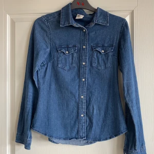 Jeansskjorta från H&M - Säljer en klassisk blå jeansskjorta från H&M med tryckknappar och två bröstfickor med lock. Skjortan har lång ärm och är tillverkad i mjukt denimtyg. Perfekt att styla till olika outfits!