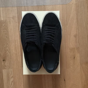 Arigato skor - Säljer ett par svarta sneakers från arigato med vit sula och diskret logga på sidan. Skorna har snörning och är tillverkade i mocka och skinn. Perfekta för en stilren och avslappnad look. Inga skador på skorna men har använt dom ett tag