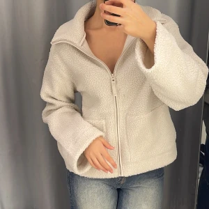 Vit teddyjacka med dragkedja - Mysig vit teddyjacka med dragkedja framtill och två fickor. Jackan har en hög krage och är långärmad med en avslappnad passform. Perfekt för kyliga dagar och enkel att matcha med jeans eller leggings.