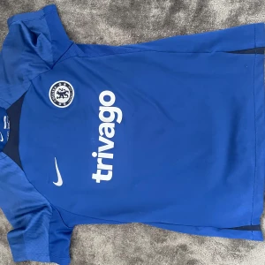 Blå Chelsea FC fotbollströja från Nike - Chelsea FC tränings t-shirt i blått med klubbmärke och Nike-logga på bröstet. Trivago sponsortryck på framsidan. Slim fit-modell med korta ärmar och ventilerande material, perfekt för fotbollsträning.