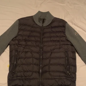 Moncler cardigan grå - Moncler cardigan grå med quiltad framsida och mjuka grå ärmar. Jackan har hög krage, dragkedja framtill och två fickor med dragkedja. Perfekt för lager-på-lager och stilren look. Priset är inte hugget i sten 