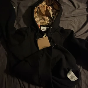Svart hoodie från Burberry med mönstrad huva - Säljer en svart hoodie från Burberry med dragkedja och klassiskt rutmönstrat foder i huvan. Tröjan har guldfärgade detaljer på snörena och en snygg, stilren look. Perfekt för dig som gillar exklusiva märken och streetstyle kom med prisförslag påse följer med .