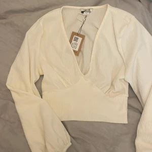 Vit v-ringad blus från NA-KD - Säljer en stilren beige/krämvit blus från Monki med djup v-ringning och långa ärmar. Toppen har en markerad midja och är tillverkad i ett lätt, strukturerat material. Perfekt för att skapa en trendig look.  fläckar på baksidan av ärmen men syns inte så mycket (bild 2)
