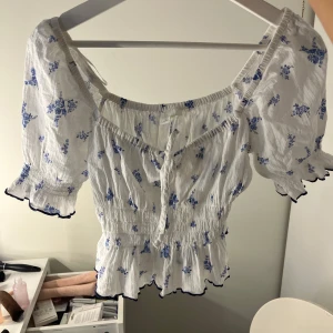 Vit blommig blus med puffärmar - Supersöt vit blus med blått blommönster och puffiga korta ärmar. Smockad midja och volangkant nertill, samt knytning fram. Perfekt för vår och sommar! 