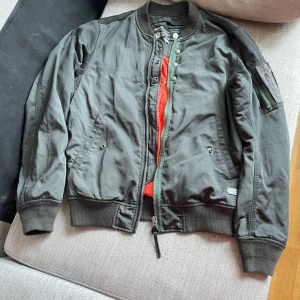 Grön bomberjacka från Diesel - Säljer en klassisk grön bomberjacka från Diesel med orange foder. Jackan har dragkedja, ribbade muddar och detaljerad patch på ärmen. Perfekt för dig som gillar streetwear och vill ha en tidlös look.