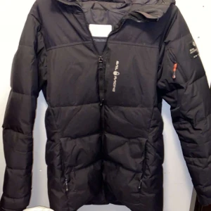 Svart dunjacka från Sail Racing - Säljer en svart dunjacka från Sail Racing med huva och dragkedja framtill. Jackan har logga på bröstet och ärmarna, samt praktiska fickor med dragkedja. Perfekt för kyliga dagar och har en sportig look.