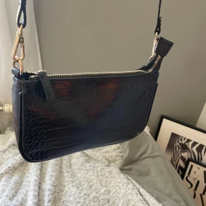 Svart axelväska med krokodilpräglat mönster - Snygg svart axelväska med krokodilpräglat mönster och guldfärgade detaljer. Väskan har dragkedja upptill och justerbar axelrem som går att ta av. Perfekt storlek för det viktigaste och stilren design som passar till många outfits.