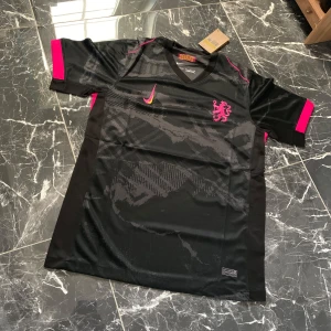 Svart och rosa fotbollströja med lejonmärke - Snygg svart fotbollströja från Nike med rosa detaljer och logga på bröstet. Tröjan har korta ärmar med rosa muddar och ett lejonmärke på vänster sida. Materialet är lätt och andas, perfekt för träning eller match.