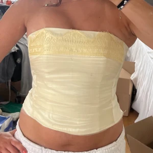 Korsett-topp - Säljer en elegant ivoryfärgad tubtopp från Lacona med vacker spetsdetalj upptill. Toppen är axelbandslös och har en glansig satinliknande yta. Stängs med hyskor baktill och har en figurnära passform.