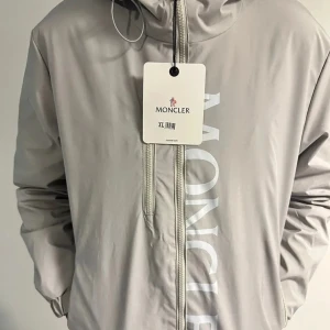 Vindjacka från Moncler - Snygg beige vindjacka från Moncler med dragkedja och huva. Jackan har Moncler-loggan tryckt vertikalt på framsidan och en liten trikolor-detalj på baksidan. Perfekt för blåsiga dagar och sommar kvällar. Jackan är helt oanvänd då den inte passa mig. 