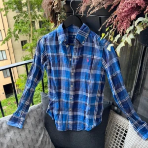 Blårutig skjorta från Ralph Lauren - Säljer en klassisk blårutig skjorta från Ralph Lauren med lång ärm och knappar framtill. Skjortan har en klassisk krage och den ikoniska rosa loggan broderad på bröstet. Perfekt för en avslappnad men stilren look.
