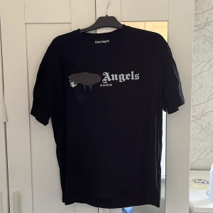 Svart t-shirt från Palm Angels - Säljer en svart t-shirt från Palm Angels med tryck på bröstet där det står 'Angels Paris' och en unik grafisk detalj över första bokstaven. T-shirten har rund hals och klassisk passform. Perfekt för dig som gillar streetwear.