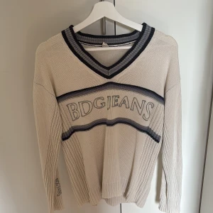 Beige stickad tröja från BDG Jeans - Säljer en beige stickad tröja från Urban Outfiters med mörkblå ränder och v-ringad hals. Tröjan har lång ärm och broderad logga framtill samt på ärmen. Perfekt för en avslappnad och stilren look.