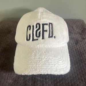 Vit keps från Classified  - Vit keps med broderad svart CLSFD-logga framtill. Framsidan är i mjuk frotté och baksidan i vit mesh för extra ventilation. Justerbar storlek med plastspänne bak. Perfekt för sommardagar. Spännet på baksidan saknar två pluppar men annars är kepsen som ny!